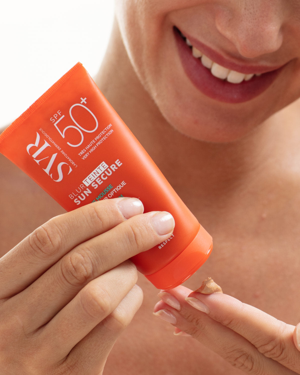 Tube orange SVR Sun Secure Blur teinté SPF50+ déposant une noisette de crème mousse teintée sur le doigt d'une femme souriante.