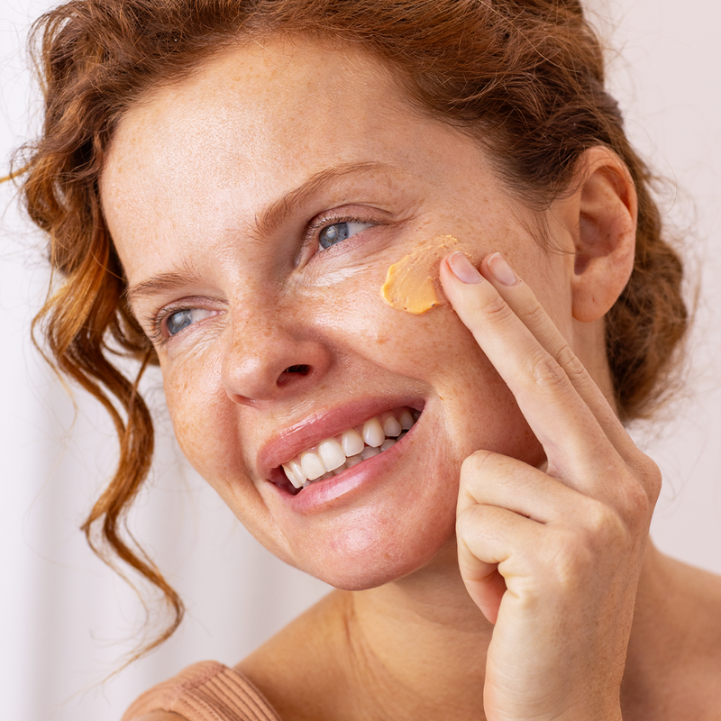 Smiling woman with red hair applying a creamy peach-colored facial mask to her cheek.