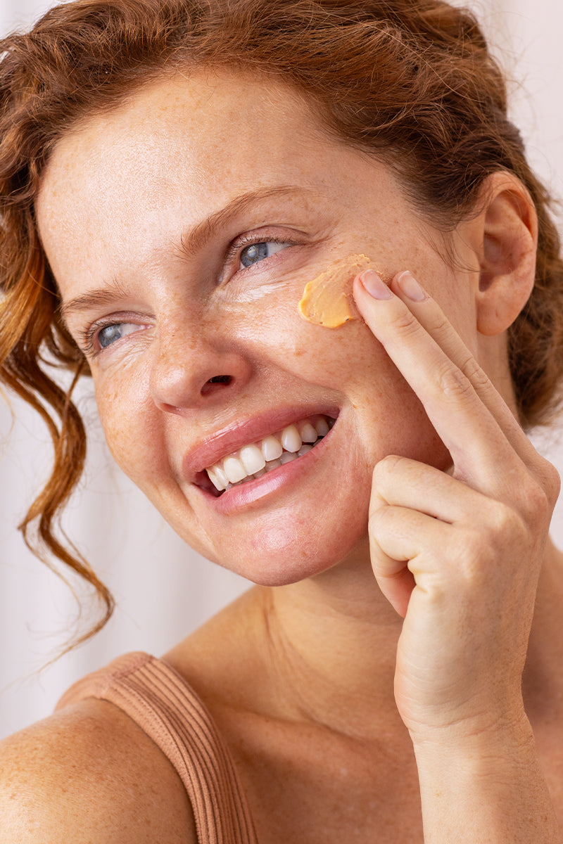 Smiling woman with red hair and freckles applying a peach-colored cream to her cheek with her finger.