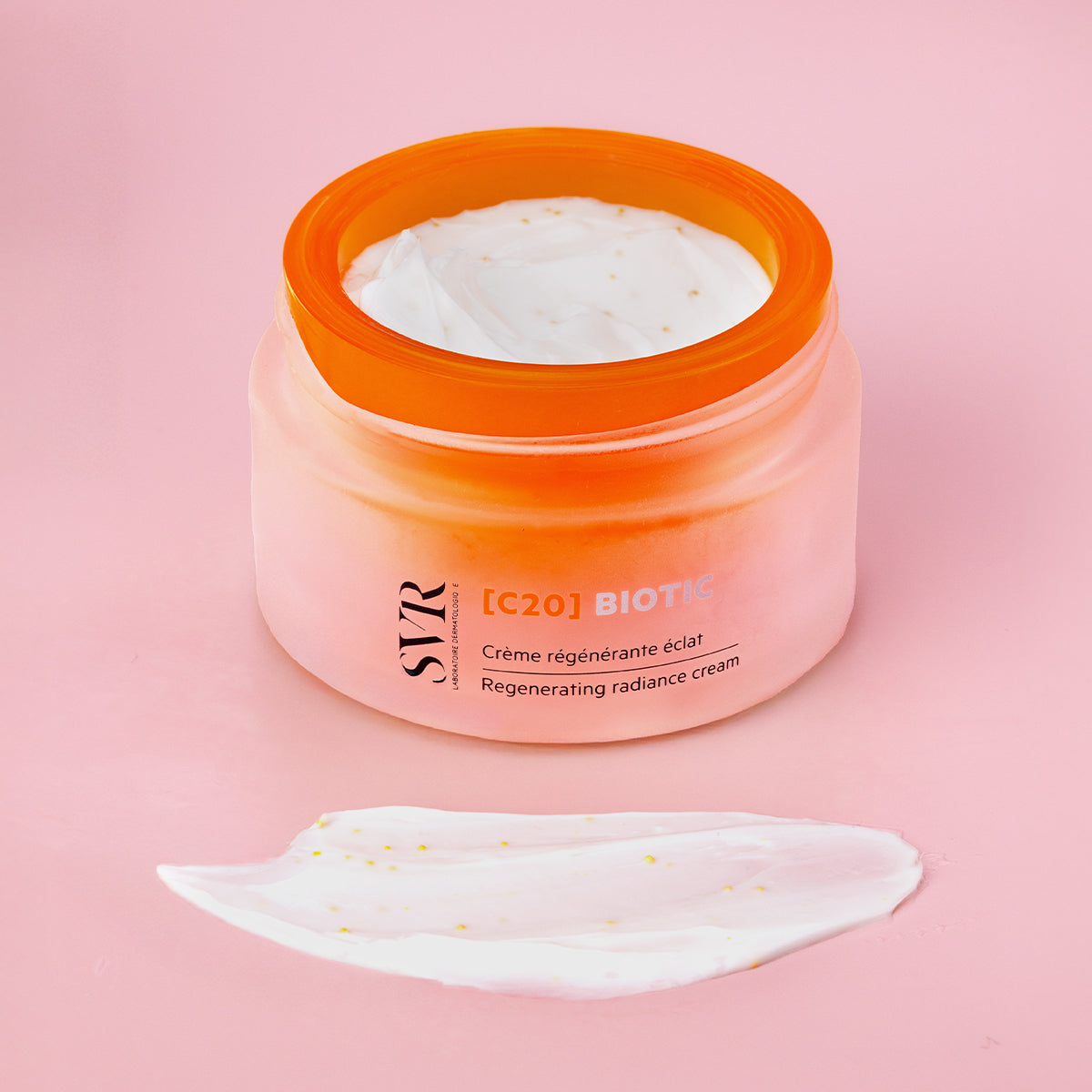 SVR [C20] Biotic regenerating radiance cream in a frosted orange jar with a white cream swatch on a pink background.