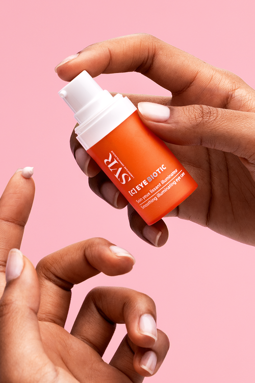Hands holding a small orange SVR [C] Eye Biotic pump bottle with a dab of white cream on a fingertip against a pink background.