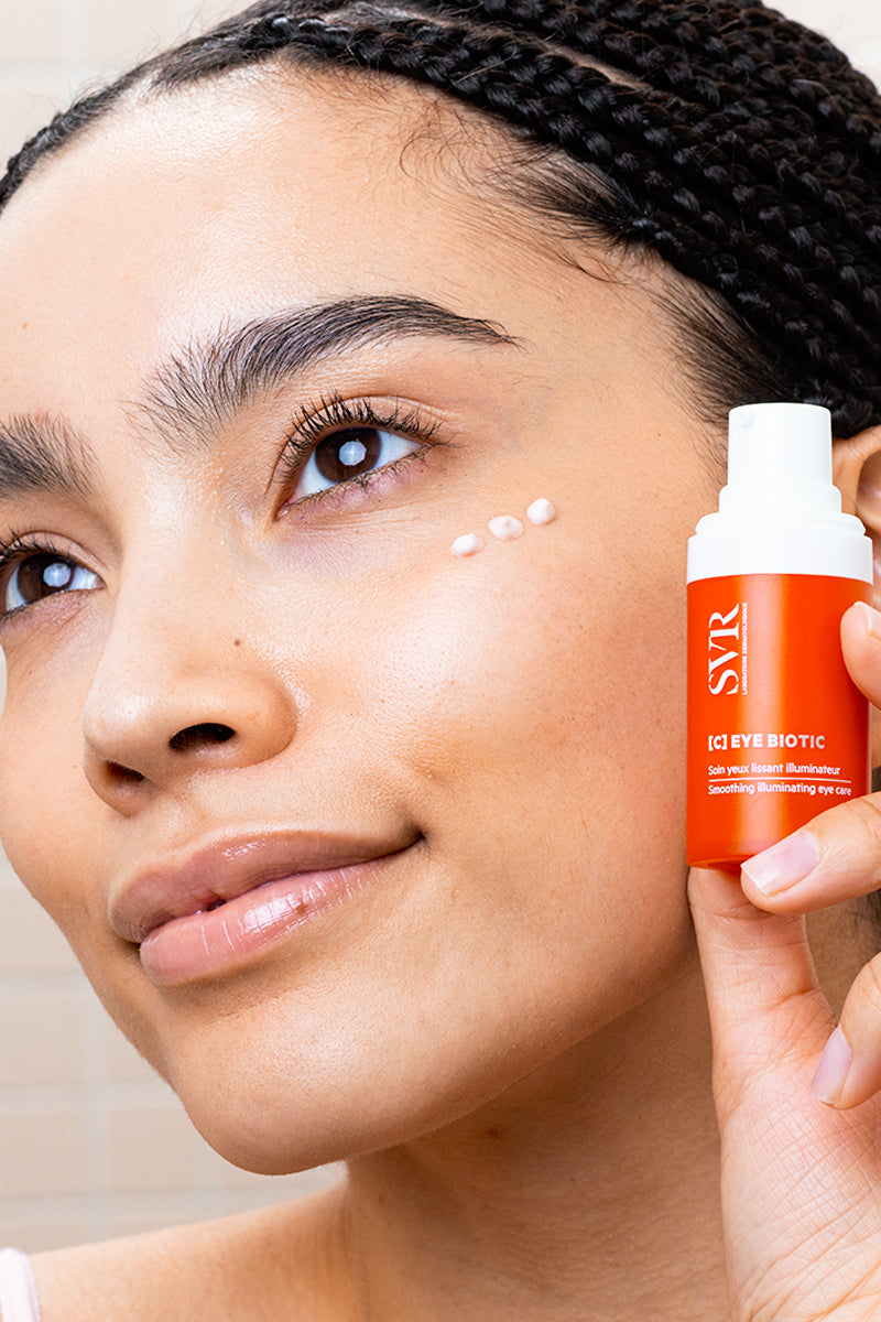 Woman holding an orange SVR [C] Eye Biotic pump bottle with cream dots applied under her eye.