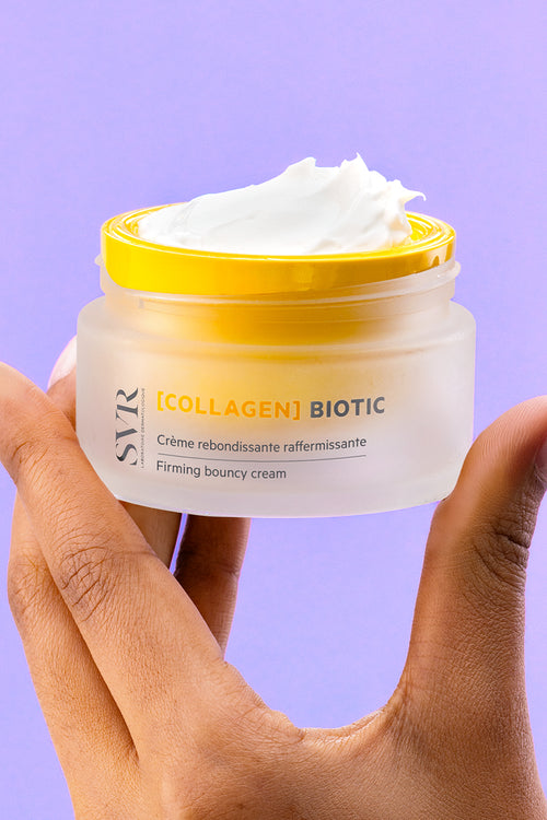 Hand holding a frosted glass jar of SVR Collagen Biotic firming cream with a bright yellow lid and white textured cream.