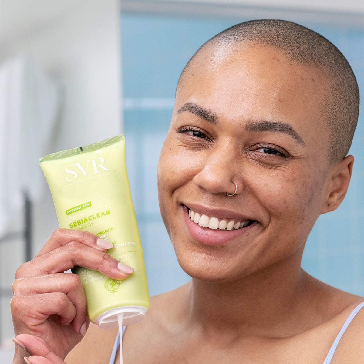 Femme souriante tenant un tube de crème lavante SVR Sebiaclear jaune pâle dans une salle de bain lumineuse.