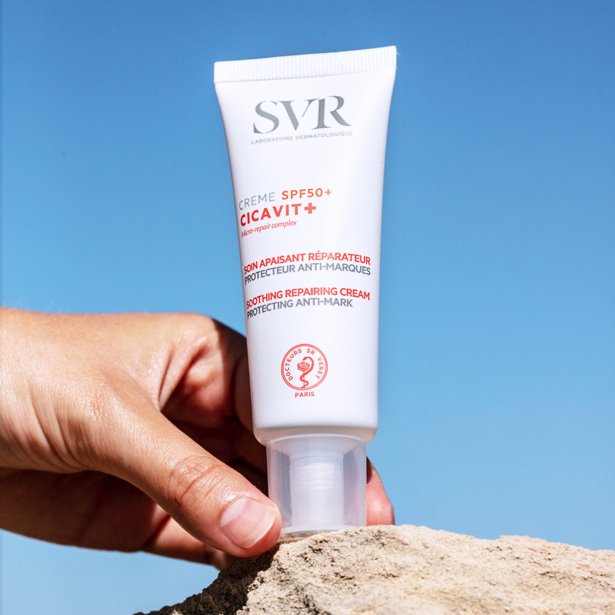 Tube blanc de crème SVR Cicavit+ SPF50+ tenu à la main sur un rocher sous un ciel bleu éclatant.