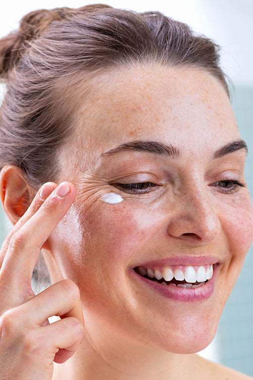 Femme souriante appliquant une noisette de crème blanche onctueuse sur le contour de ses yeux sensibles et irrités.