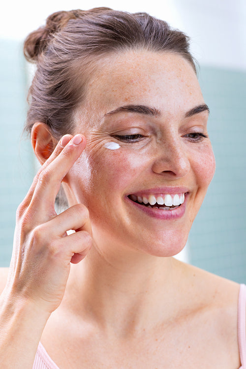 Smiling woman with freckles gently applying white moisturizing cream under her eye in a bright bathroom.