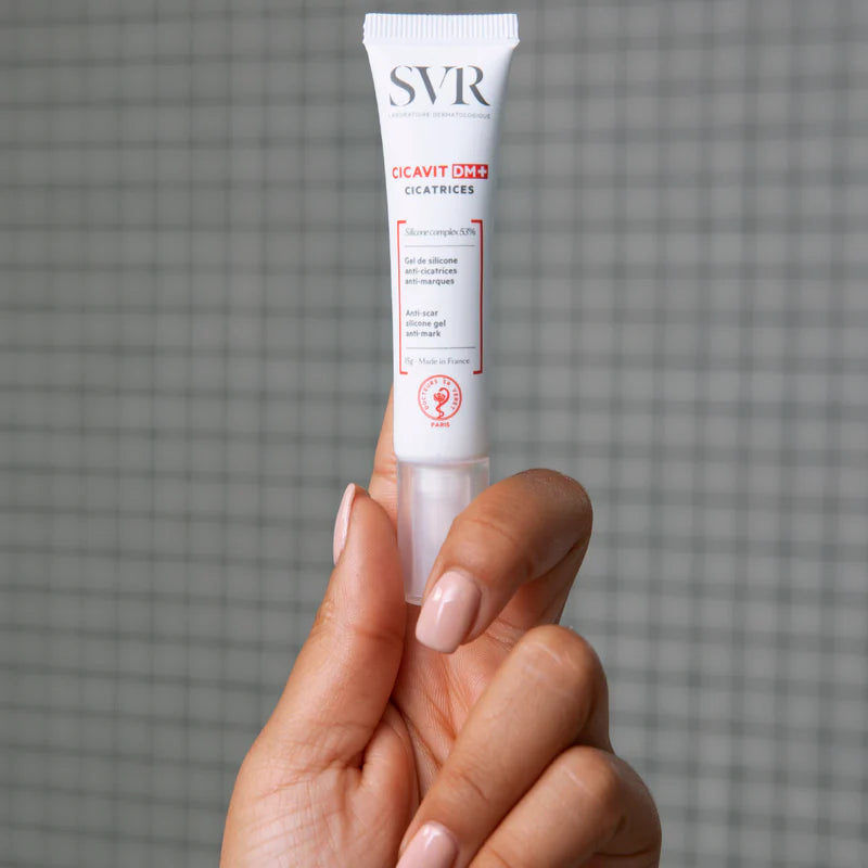 Tube blanc de gel cicatrisant SVR Cicavit DM+ de 15g tenu par une main sur fond carrelé gris.