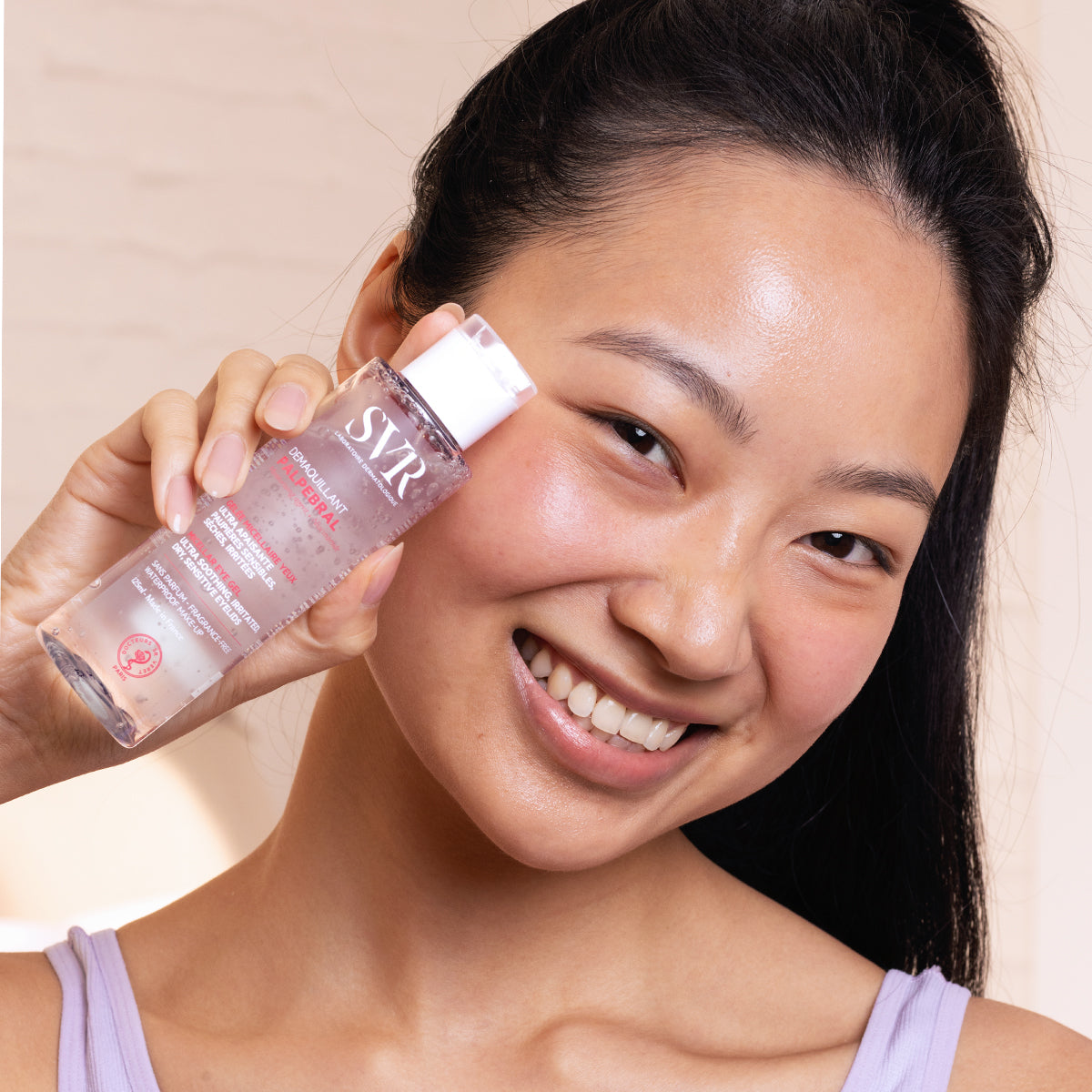 Femme souriante tenant un flacon transparent de démaquillant SVR Palpebral à la texture gelée pour yeux sensibles.