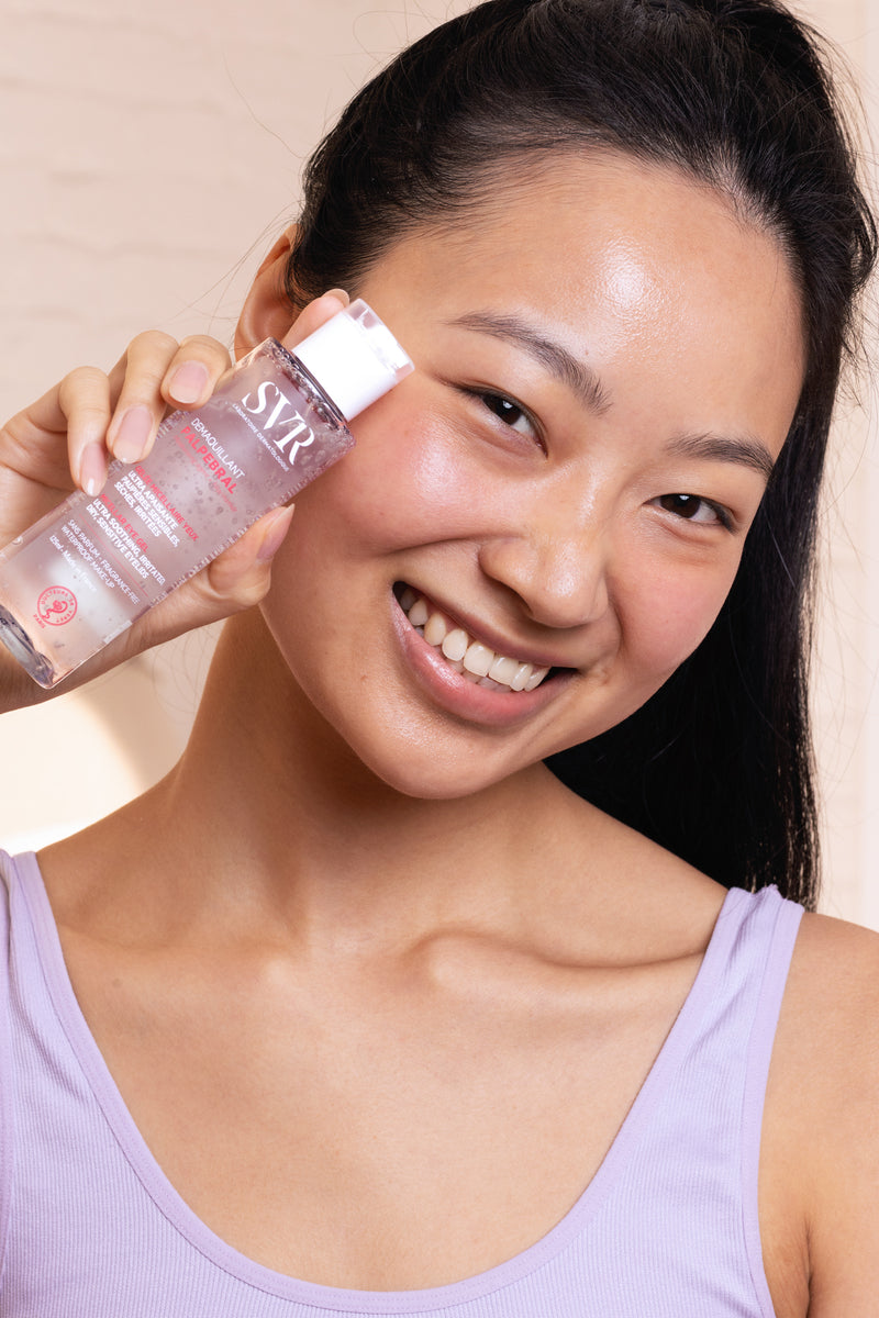 Smiling woman holding a clear bottle of SVR Palpebral makeup remover against her cheek.