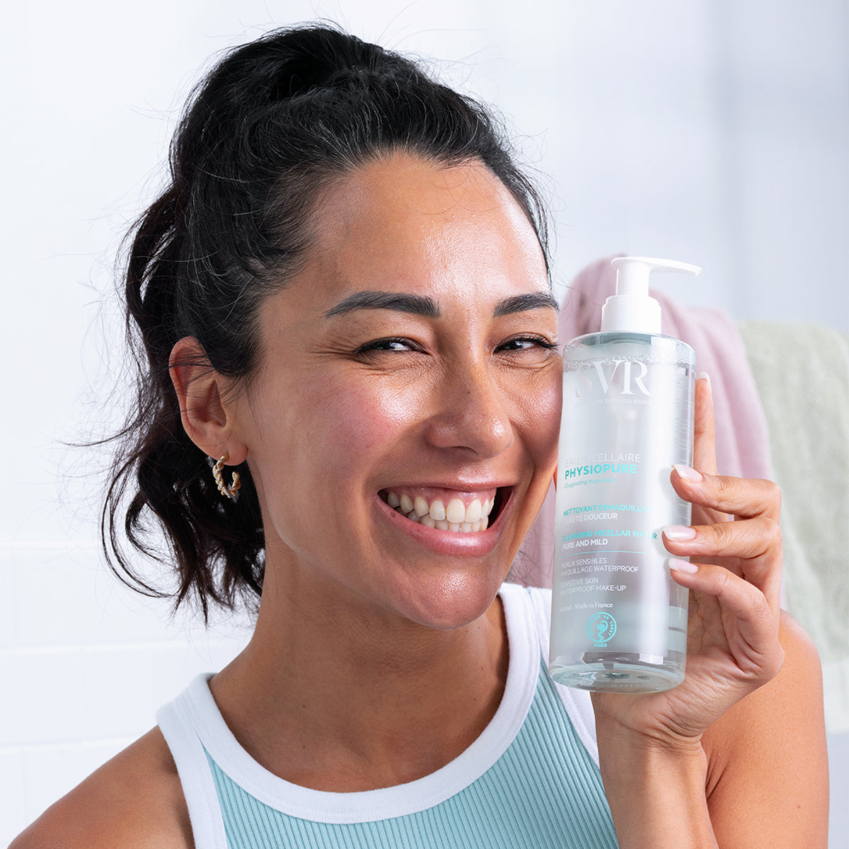 Femme souriante tenant un flacon pompe transparent de SVR PHYSIOPURE Eau Micellaire pour peaux sensibles.