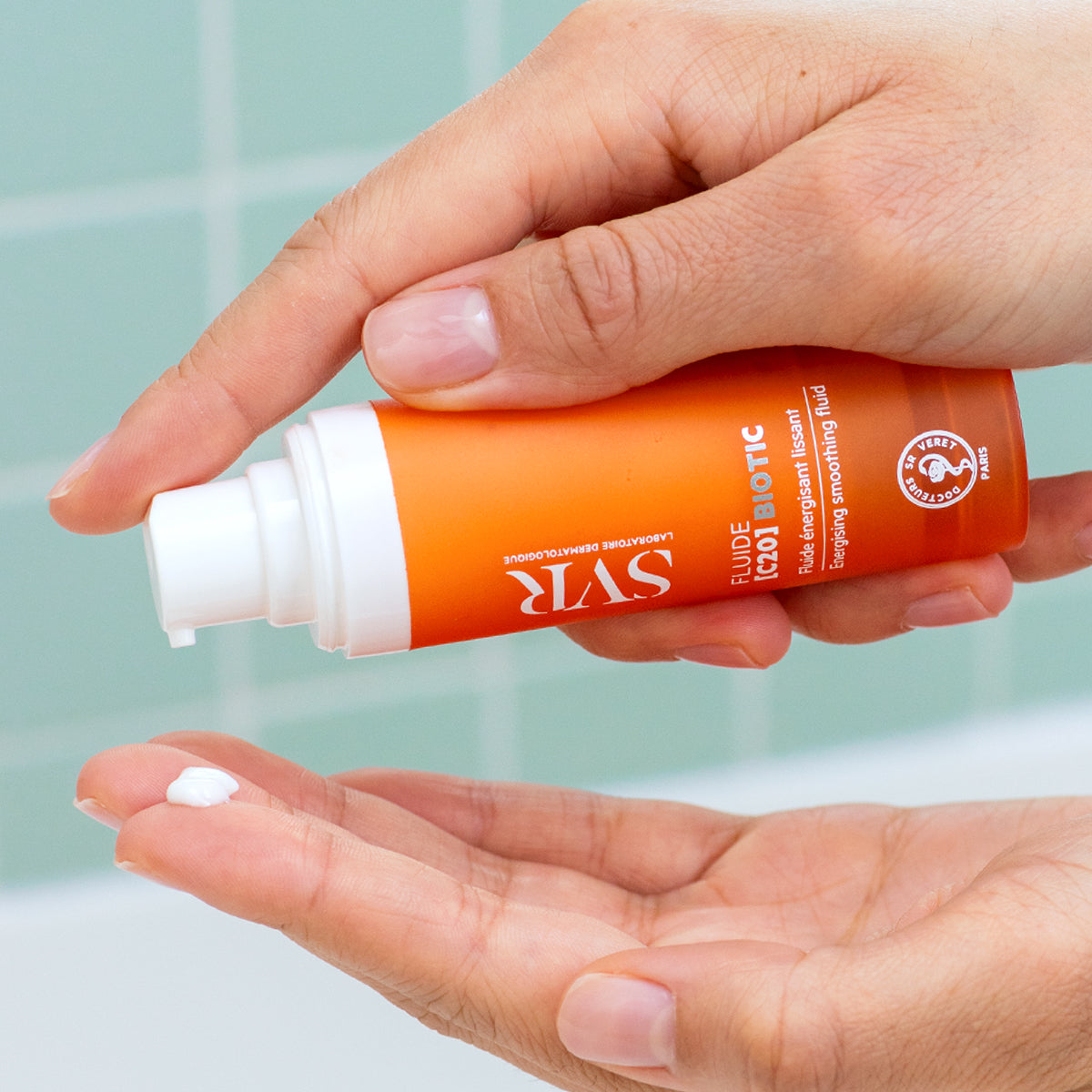Hand dispensing white SVR Fluide [C20] BIOTIC energizing smoothing fluid from an orange pump bottle.