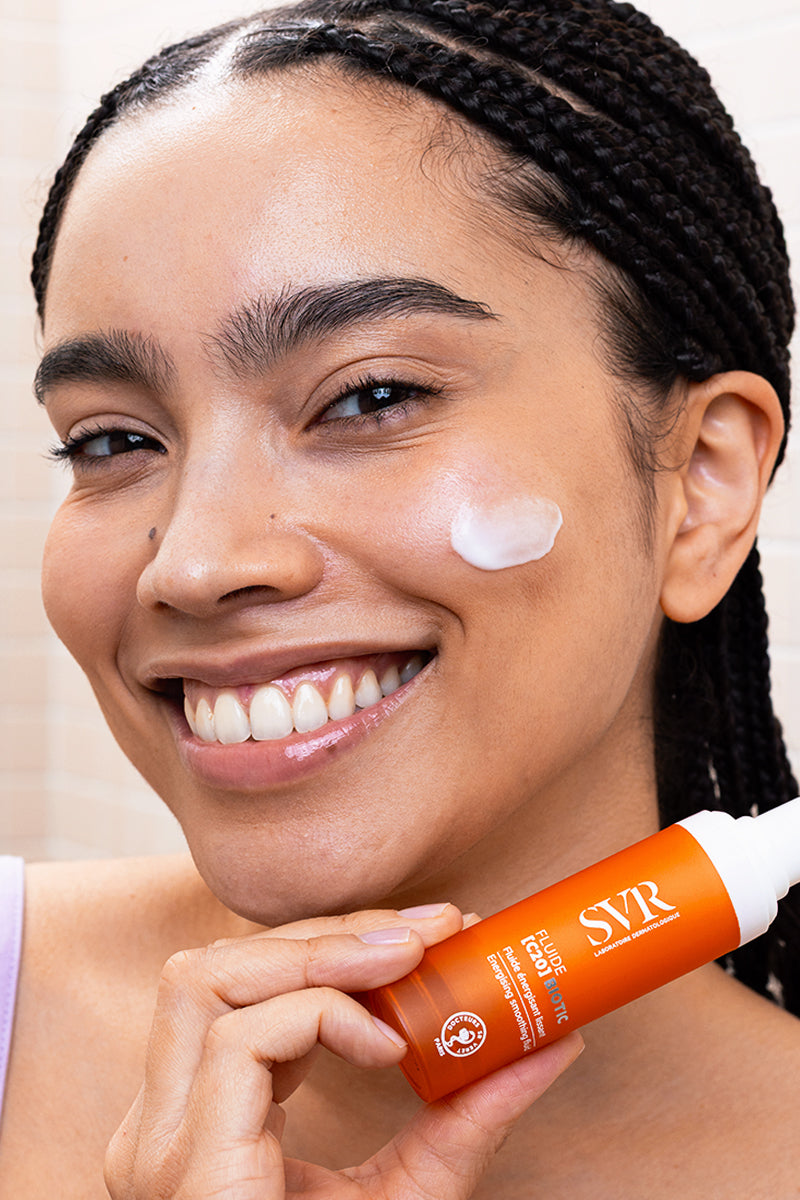 Smiling woman holding an orange SVR Fluide [C20] BIOTIC pump bottle with cream applied to her cheek.