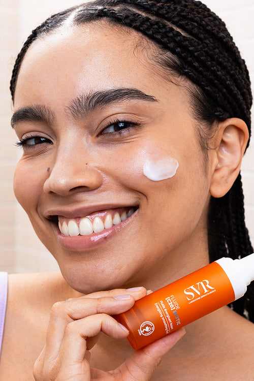 Smiling woman holding an orange SVR Fluide [C20] BIOTIC pump bottle with cream applied to her cheek.