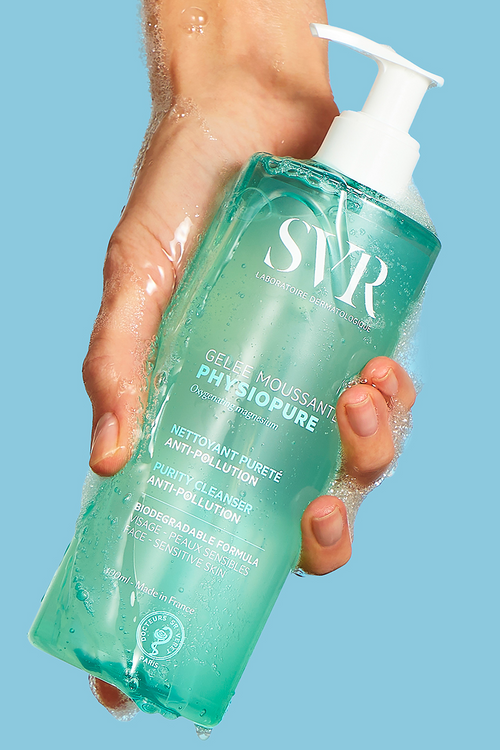 Main tenue en main d'un flacon pompe SVR Physiopure Gelée Moussante bleu translucide avec une mousse nettoyante légère.