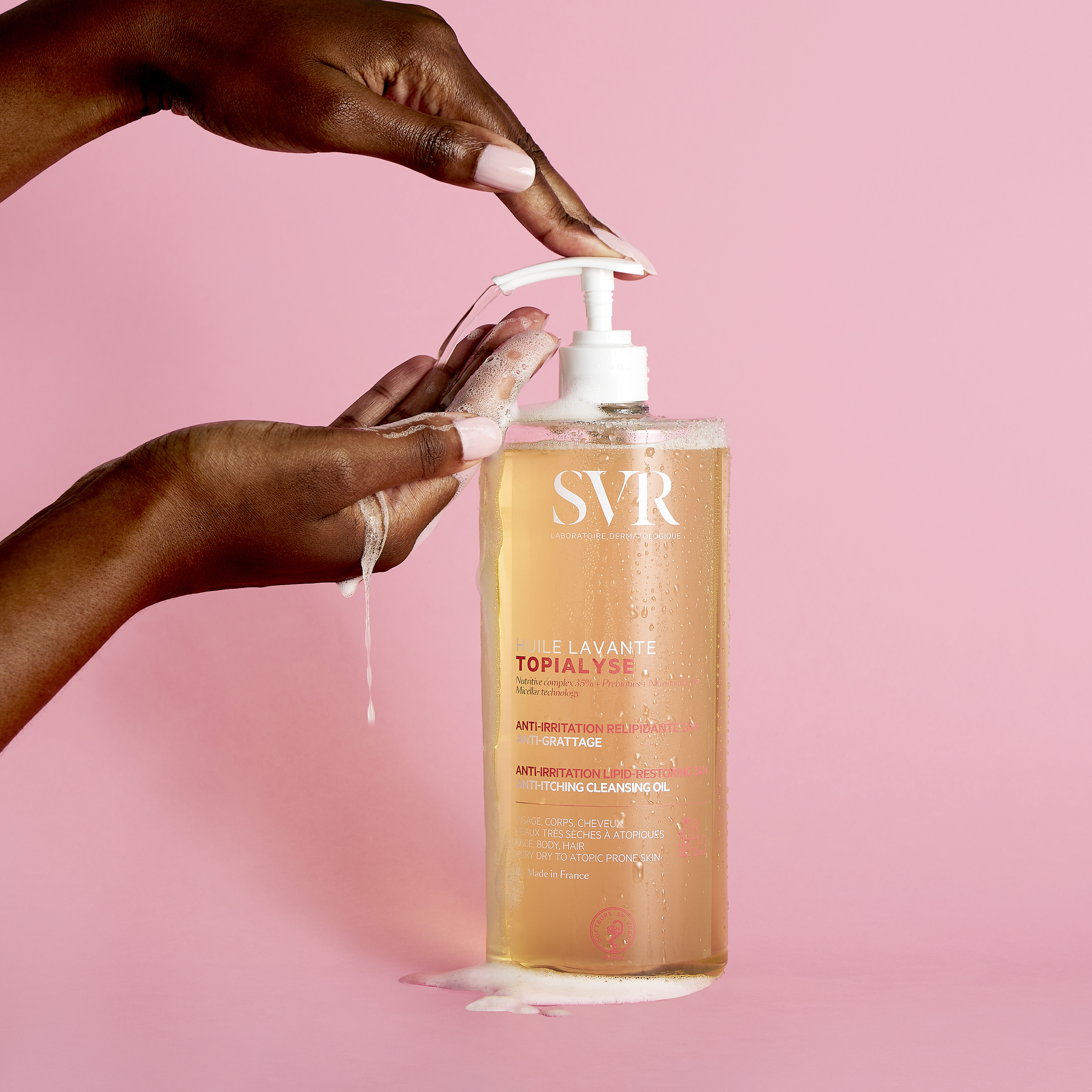 Hands dispensing SVR Topialyse cleansing oil from a large pump bottle against a soft pink background.