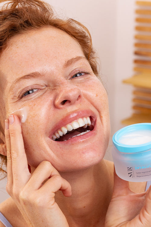 Smiling woman applying SVR HYALU Biotic regenerating plumping gel from a frosted glass jar with a blue lid.