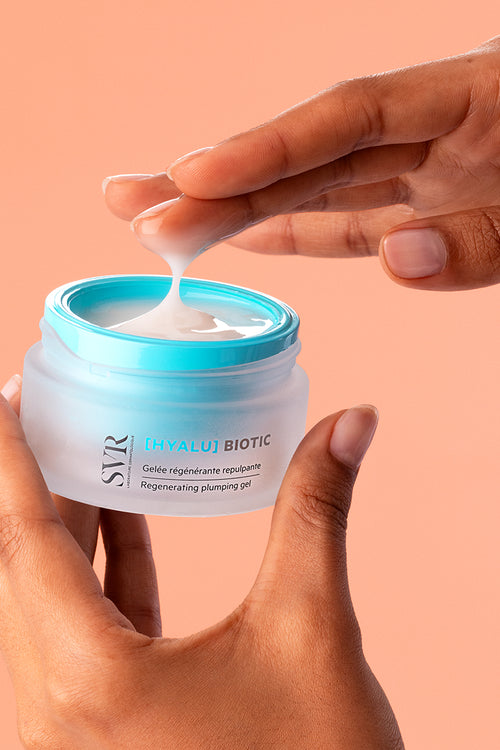 Hands holding a frosted glass jar of SVR Hyalu Biotic regenerating plumping gel with a bright blue lid and white cream.