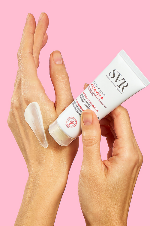 SVR CICAVIT+ HPPI cream tube held in hands with a smooth white texture applied to the skin against a pink background.