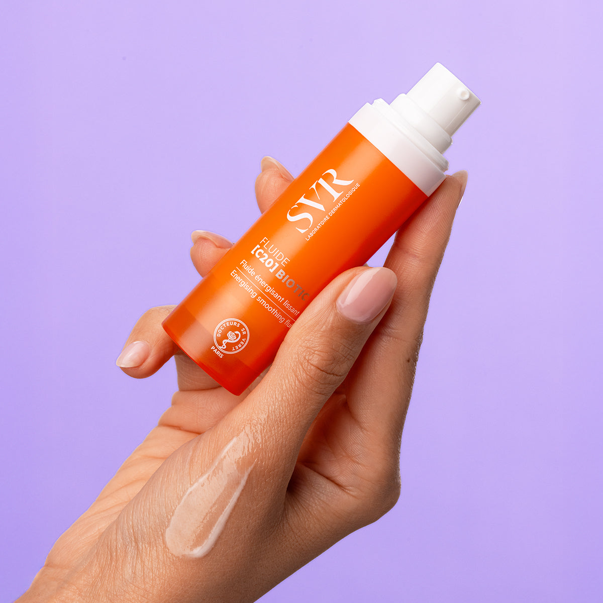 Hand holding an orange SVR Fluide [C20] BIOTIC pump bottle with a clear serum swatch against a purple background.