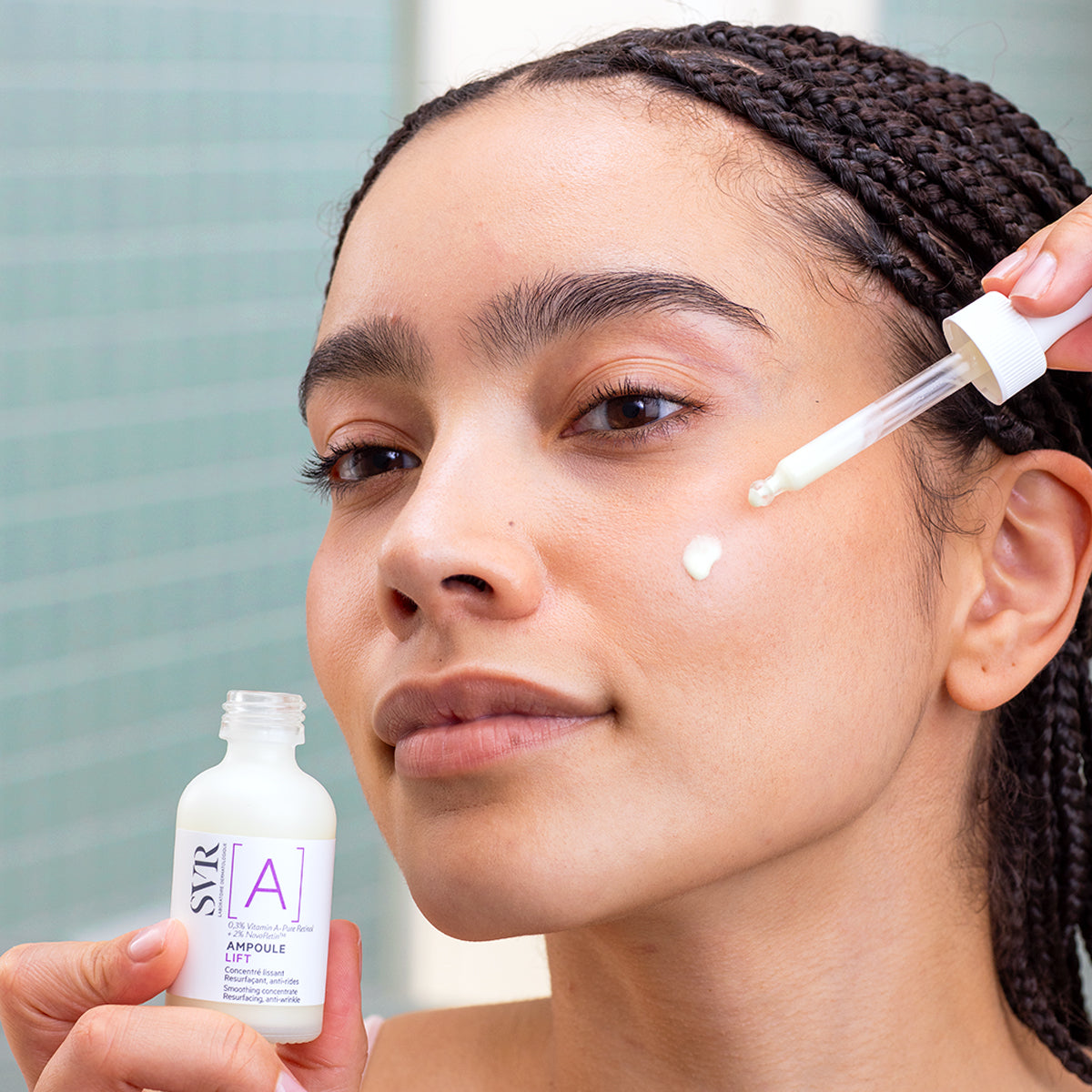 Woman applying SVR Ampoule Lift serum with a glass dropper onto her cheek from a small white bottle.