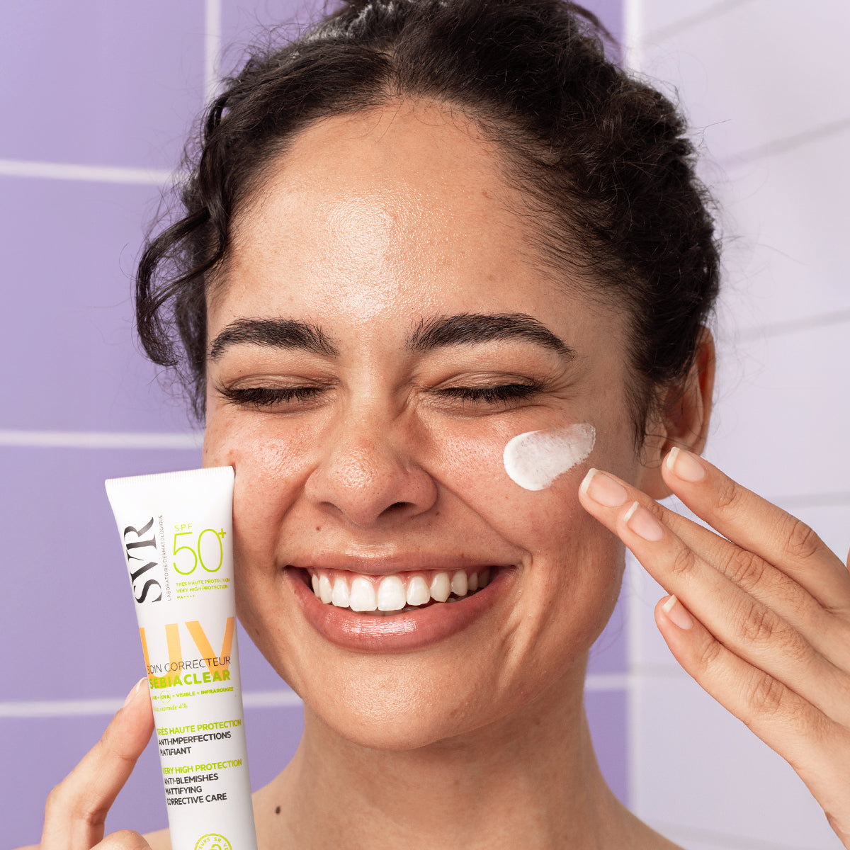 Femme souriante appliquant la crème SVR Sebiaclear SPF50+ sur sa joue devant un carrelage violet.