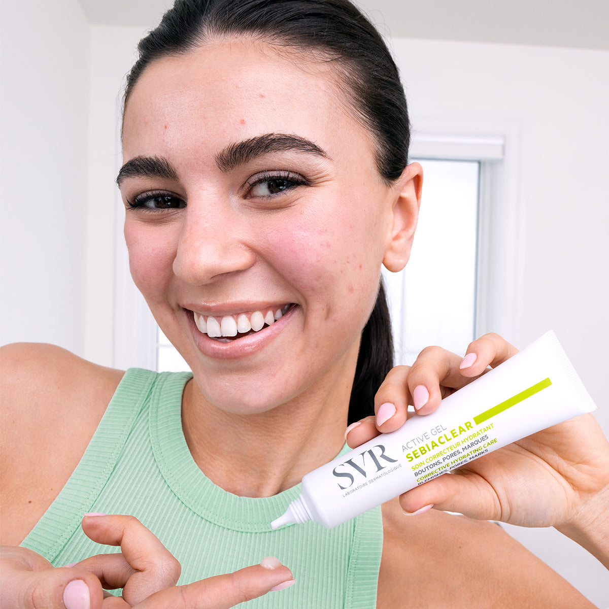 Smiling woman holding a white tube of SVR Sebiaclear Active Gel with a drop of clear product on her fingertip.