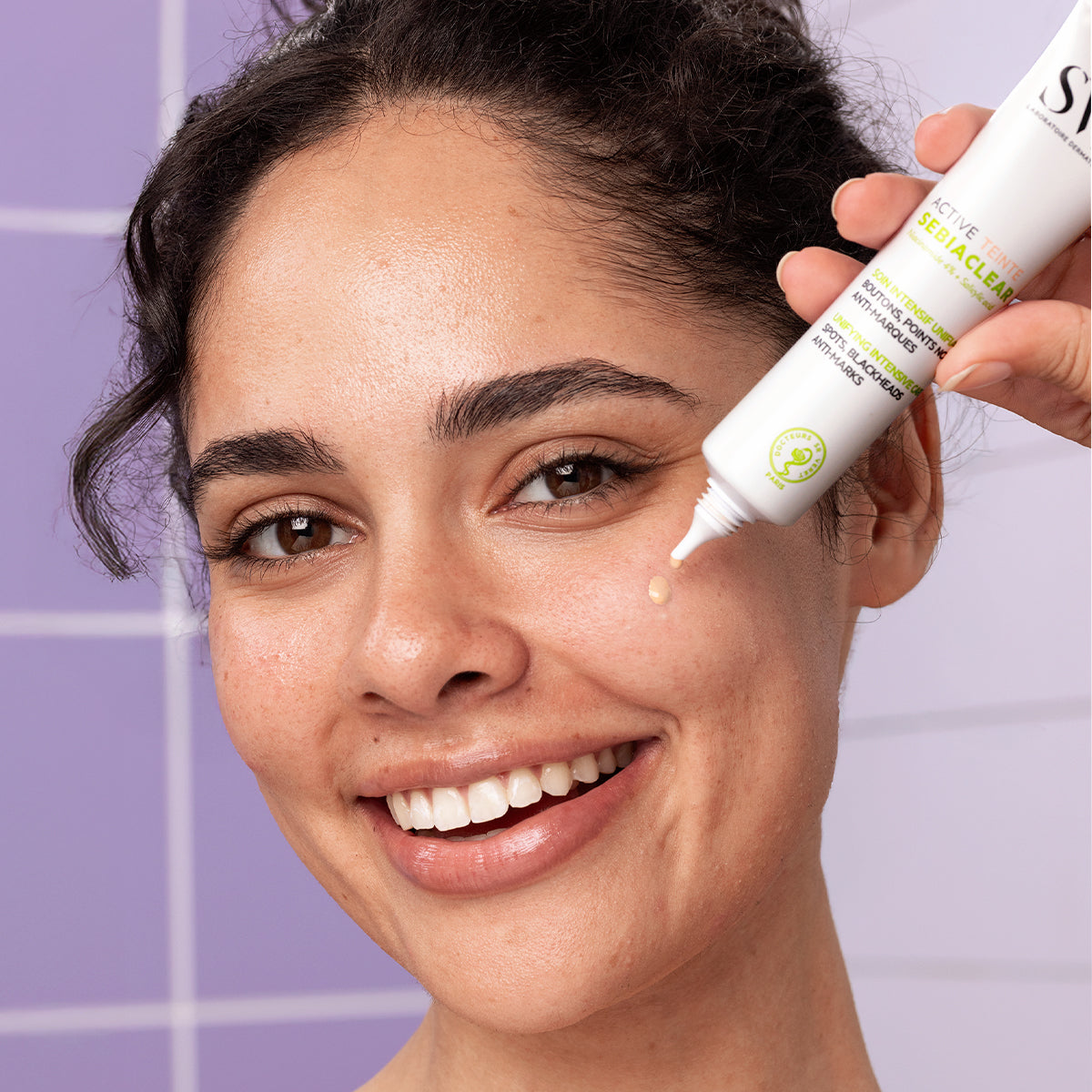 Smiling woman applying a drop of white SEBIACLEAR Active Teinté cream from a squeeze tube onto her cheek.