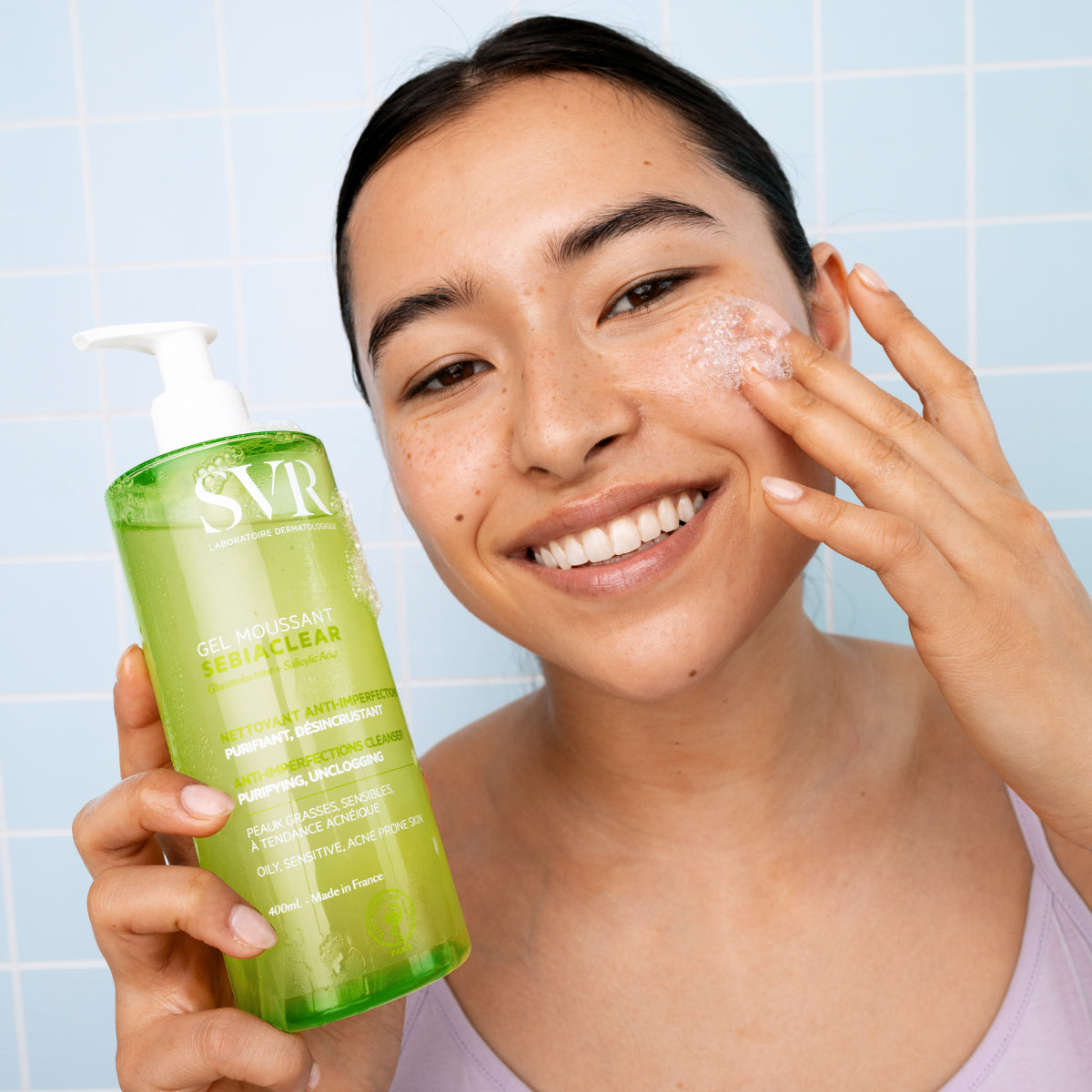 Femme souriante utilisant le gel moussant SVR Sebiaclear vert dans un flacon pompe de 400ml sur fond de carrelage bleu.