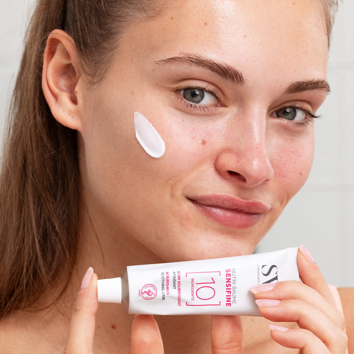 Femme appliquant le baume hydratant SVR Sensifine Nutri-Baume sur sa joue avec le tube en aluminium en main.