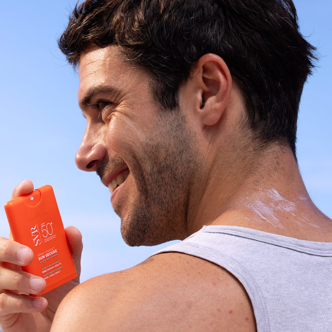 Man holding a bright orange SVR SUN SECURE SPF50+ pocket spray bottle against a clear blue sky.